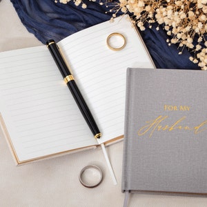 May include: Two gray journals with gold accents. One journal has the words "For My Husband" written in gold script. The other journal is open with a black pen on top of the pages.