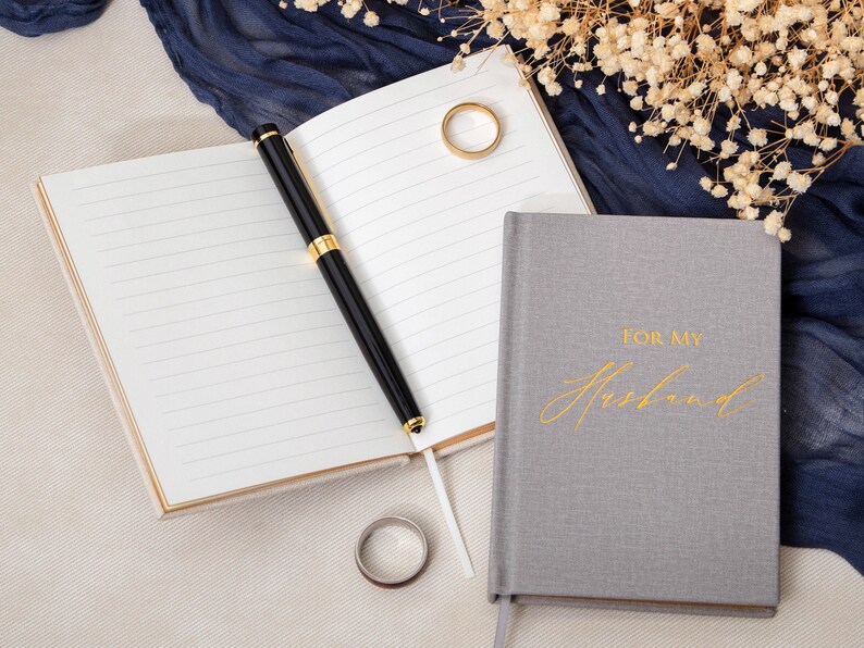 May include: Two gray journals with gold accents. One journal has the words "For My Husband" written in gold script. The other journal is open with a black pen on top of the pages.