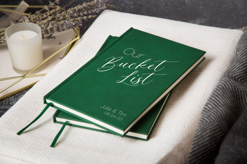 Puede incluir: Un diario de tapa dura verde con el texto "Our Bucket List" en letras blancas cursivas. El diario tiene un marcador de cinta blanco y los nombres "Julia & Tim" y la fecha "08.24.23" est&aacute;n impresos en la parte inferior.