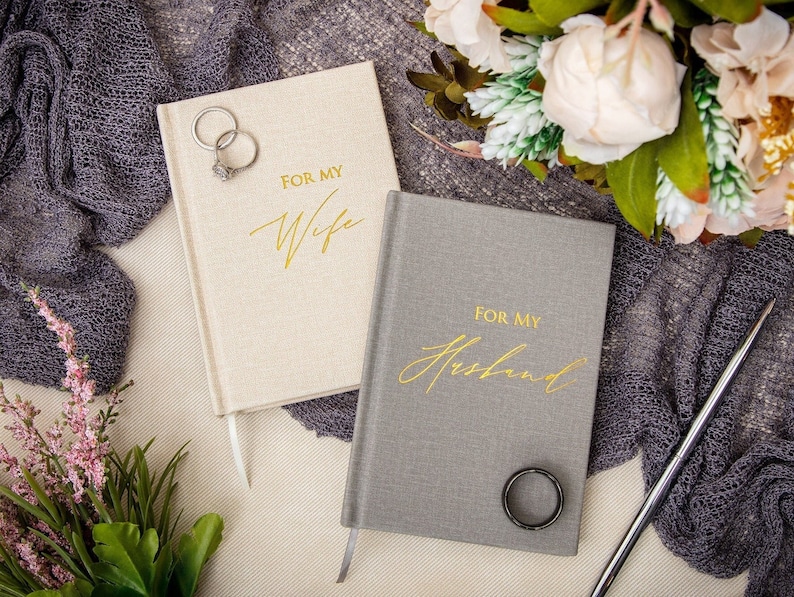 May include: Two journals with linen covers, one beige and one gray. The covers have gold lettering that reads "For My Wife" and "For My Husband".