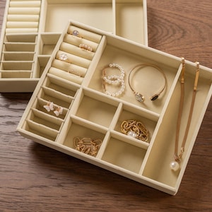 May include: Beige jewellery organiser with multiple compartments and ring slots. The organiser holds various jewellery pieces, including rings, bracelets, necklaces, and earrings. The jewellery is gold and pearl-coloured.