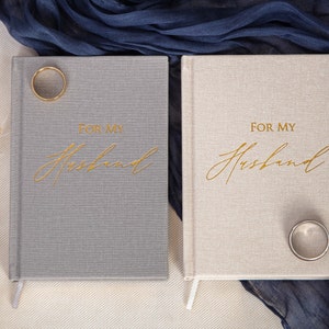 May include: Two gray and beige linen-covered journals with gold lettering that reads "For My Husband". A gold ring is on top of each journal.