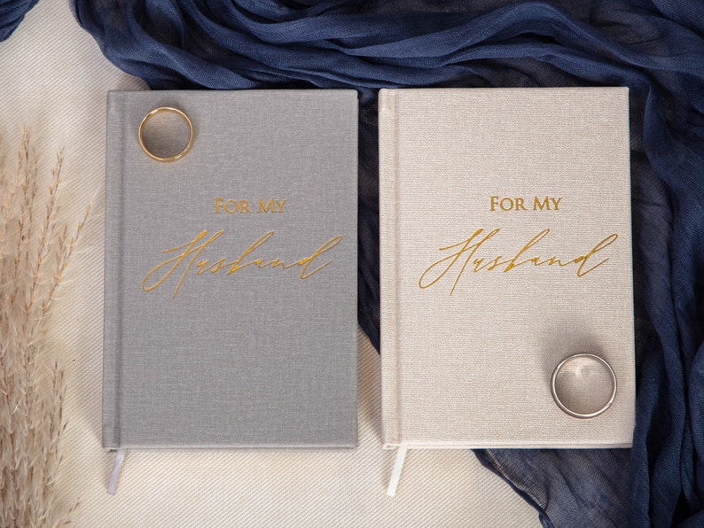 May include: Two gray and beige linen-covered journals with gold lettering that reads "For My Husband". A gold ring is on top of each journal.