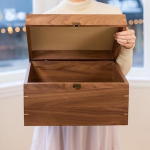 May include: A rectangular wooden box crafted from dark brown walnut wood with a hinged lid. The box features light-colored wood inlay accents at the corners and a small, metallic bronze-toned latch centered on the front edge of the lid and base.