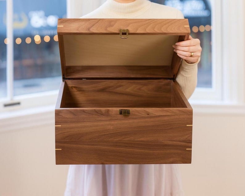 May include: A rectangular wooden box crafted from dark brown walnut wood with a hinged lid. The box features light-colored wood inlay accents at the corners and a small, metallic bronze-toned latch centered on the front edge of the lid and base.