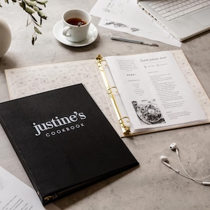 May include: A black cookbook titled "Justine's Cookbook" with white lettering, open to a recipe page. A cup of tea, a laptop, and a pair of white earbuds are also on the table. The cookbook has gold-colored binder rings.