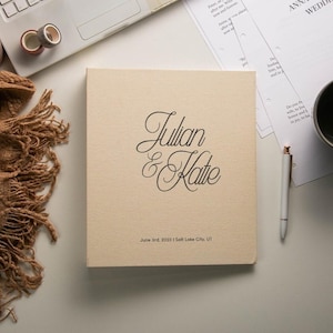 May include: A square, beige, fabric-covered wedding album featuring the black script text Julian & Katie, with the date June 3rd, 2023 and the location Salt Lake City, UT printed in a smaller, simple black font at the bottom center.
