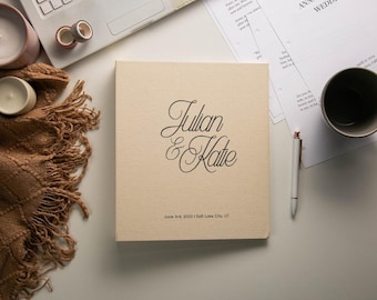 May include: A square, beige, fabric-covered wedding album featuring the black script text Julian & Katie, with the date June 3rd, 2023 and the location Salt Lake City, UT printed in a smaller, simple black font at the bottom center.