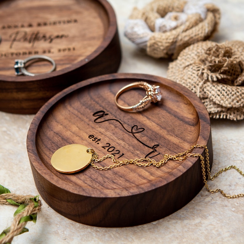 May include: Two wooden ring dishes with engraved text. The larger dish says "And they lived happily ever after" and the smaller dish says "est. 2021". A gold ring and necklace are on the smaller dish.