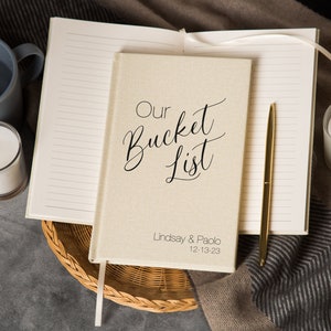 Puede incluir: Un diario beige cubierto de lino con el texto "Our Bucket List" en escritura cursiva negra. El diario est&aacute; abierto para revelar p&aacute;ginas rayadas y un bol&iacute;grafo dorado. Los nombres "Lindsay & Paolo" y la fecha "12-13-23" est&aacute;n impresos en la portada.