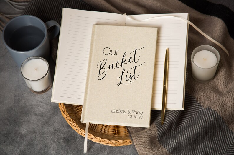 Puede incluir: Un diario beige cubierto de lino con el texto "Our Bucket List" en escritura cursiva negra. El diario est&aacute; abierto para revelar p&aacute;ginas rayadas y un bol&iacute;grafo dorado. Los nombres "Lindsay & Paolo" y la fecha "12-13-23" est&aacute;n impresos en la portada.