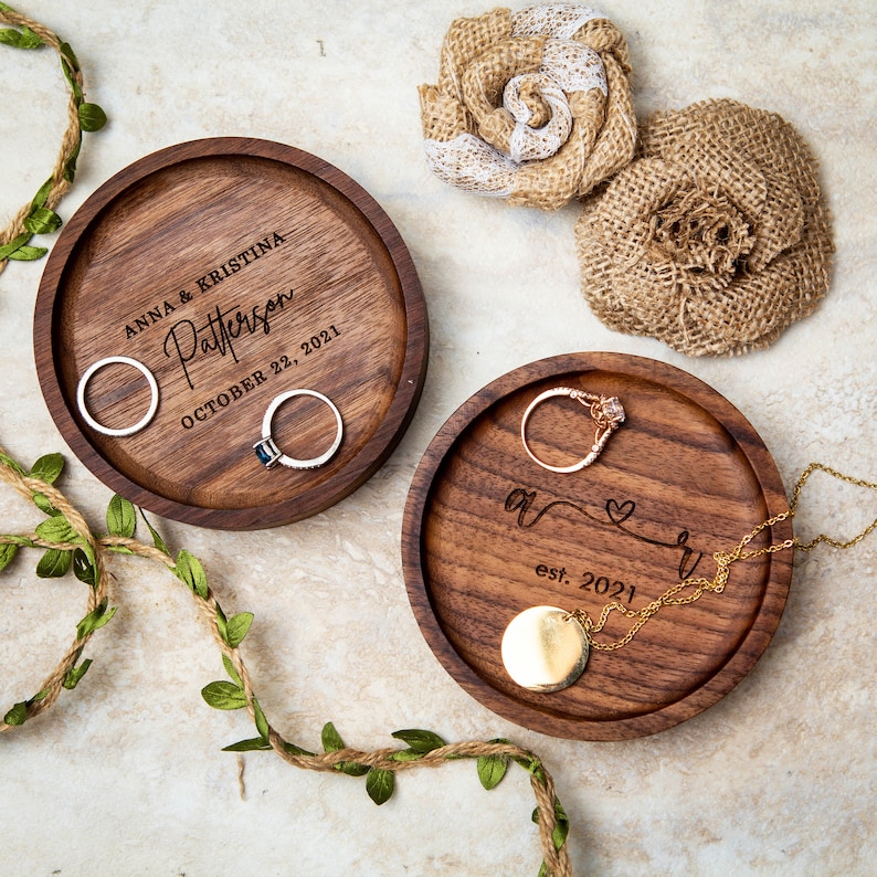 May include: Two wooden ring dishes with engraved text. The first dish says "Anna & Kristina Patterson October 22, 2021". The second dish says "A & R est. 2021" with a heart symbol.