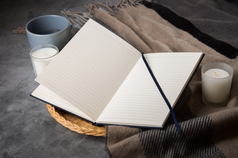 Puede incluir: Un cuaderno abierto con l&iacute;neas y un marcador de cinta azul en una cesta tejida, junto a dos velas blancas y una taza gris sobre una superficie gris.