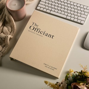May include: A beige binder with the words "The Officiant" and the name "Michael Thomas" printed in black. The names "Emily & Harry" and the date "09.09.2023" are printed on the bottom. A white keyboard and mouse are in the background.