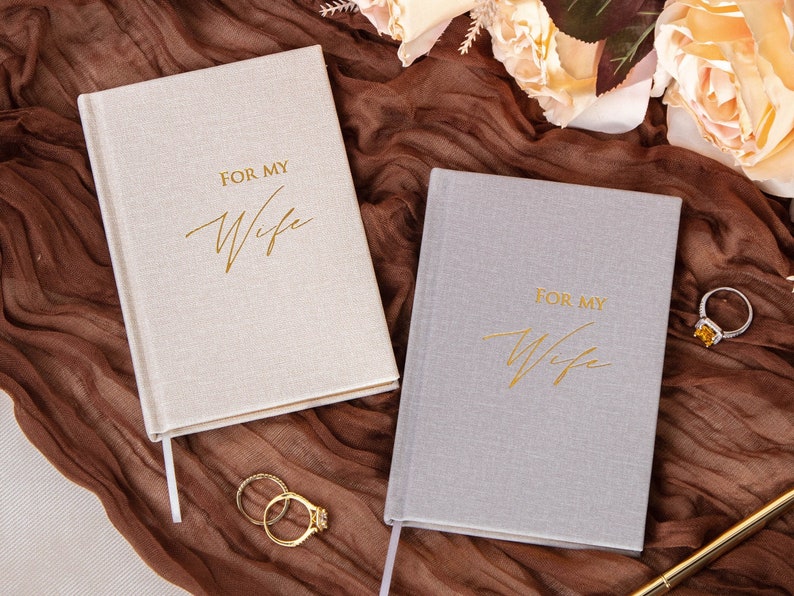 May include: Two gray journals with gold lettering that reads "For My Wife". The journals are on a brown fabric background with two gold rings.