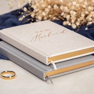 May include: Two journals with gold trim and linen covers. The top journal has the text "To My Husband" in gold lettering. A gold wedding band is in the foreground.