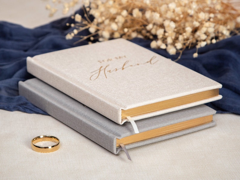 May include: Two journals with gold trim and linen covers. The top journal has the text "To My Husband" in gold lettering. A gold wedding band is in the foreground.