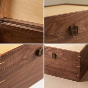 May include: A rectangular wooden box crafted from dark brown walnut wood with a lighter wood lid. The box features visible corner joinery with light-colored wood splines, a metal latch closure, and metal hinges on the interior lid attachment.