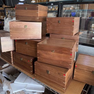 May include: A collection of rectangular wooden boxes made from dark walnut wood. Each box features a hinged lid, a metallic latch closure on the front, and visible light-colored wood joinery details at the corners of the lid and base.