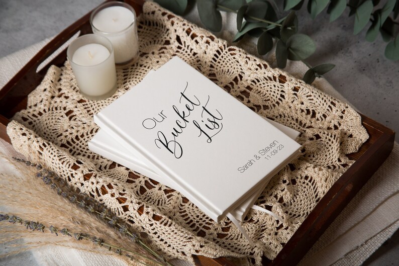 Puede incluir: Un libro blanco con el texto "Our Bucket List" en caligraf&iacute;a negra. El libro est&aacute; personalizado con los nombres "Sarah & Steven" y la fecha "11.08.22".