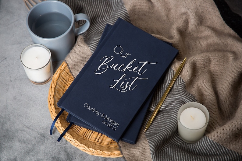 Puede incluir: Un diario azul marino con el texto "Our Bucket List" en escritura blanca. El diario est&aacute; personalizado con los nombres "Courtney & Morgan" y la fecha "08.30.22".