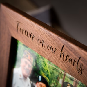Op de afbeelding: Een houten fotolijst met een foto van twee mensen in uniform. De lijst is gegraveerd met de woorden "Forever in our hearts".