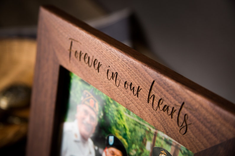 Op de afbeelding: Een houten fotolijst met een foto van twee mensen in uniform. De lijst is gegraveerd met de woorden "Forever in our hearts".