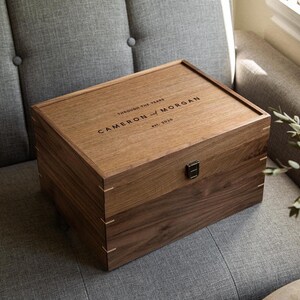 May include: A rectangular wooden keepsake box with a dark brown finish and a hinged lid. The lid is engraved with the text "THROUGH THE YEARS", "CAMERON AND MORGAN", and "EST. 2020". A small black metal latch is centered on the front of the box.