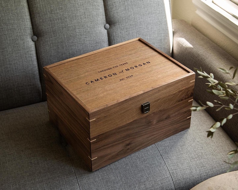 May include: A rectangular wooden keepsake box with a dark brown finish and a hinged lid. The lid is engraved with the text "THROUGH THE YEARS", "CAMERON AND MORGAN", and "EST. 2020". A small black metal latch is centered on the front of the box.