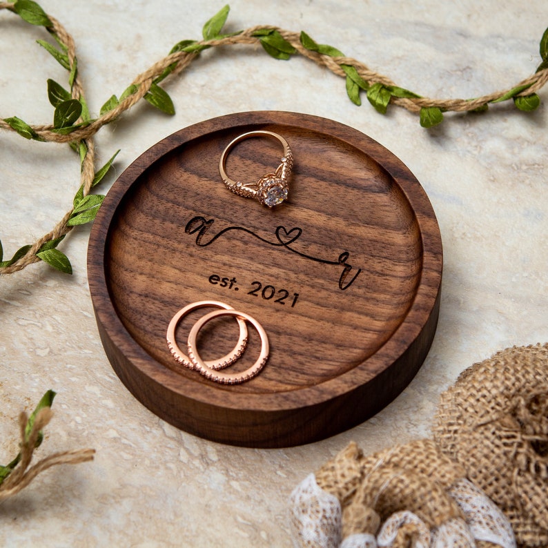 May include: A round wooden ring dish with a dark brown finish. The dish has a personalized inscription that reads "A & R est. 2021" with a heart symbol. Two gold rings are resting on the dish.