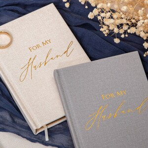 May include: Two gray and beige journals with gold lettering that reads "For My Husband". The journals are on a blue fabric background with white flowers.