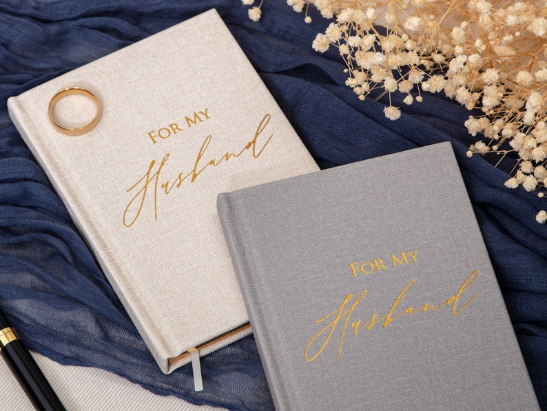 May include: Two gray and beige journals with gold lettering that reads "For My Husband". The journals are on a blue fabric background with white flowers.