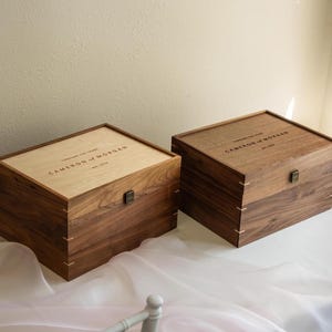 May include: Two rectangular wooden keepsake boxes with hinged lids and metal latches. One box features a light-colored wood lid, while the other is entirely dark wood. Both are engraved with the text Cameron and Morgan, est. 2019.