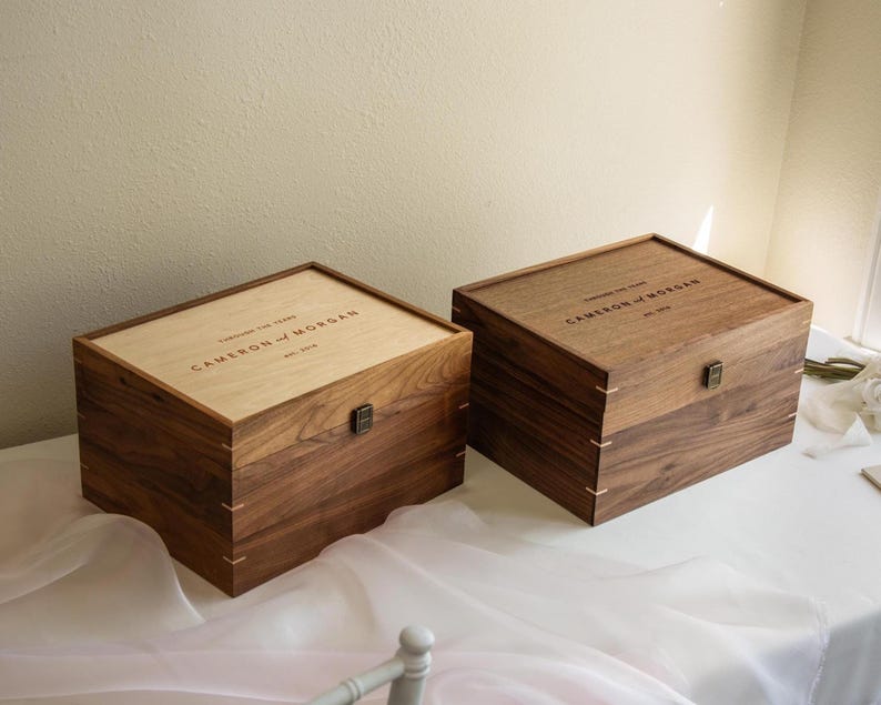 May include: Two rectangular wooden keepsake boxes with hinged lids and metal latches. One box features a light-colored wood lid, while the other is entirely dark wood. Both are engraved with the text Cameron and Morgan, est. 2019.