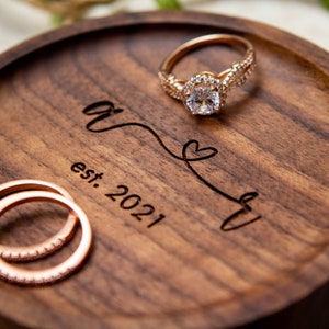 May include: A wooden ring dish with the inscription "&" and "est. 2021" with a heart symbol. Two gold rings with small diamonds are on the dish.