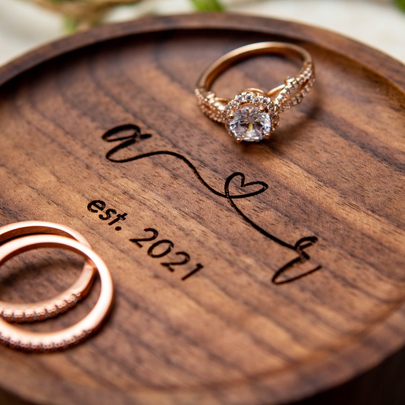 May include: A wooden ring dish with the inscription "&" and "est. 2021" with a heart symbol. Two gold rings with small diamonds are on the dish.
