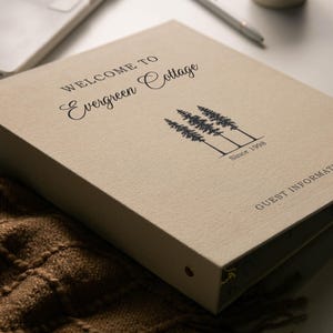 May include: A beige three-ring binder with a textured fabric cover. The front features black printed text that reads Welcome to Evergreen Cottage, a graphic of three pine trees, the text Since 1998, and the words Guest Information.