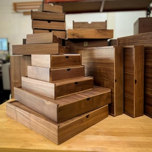 May include: A collection of various sized wooden boxes and drawers, stacked and arranged on a wooden surface. The boxes are made of a rich, dark brown wood, with visible wood grain. Some boxes have small, circular finger pulls.