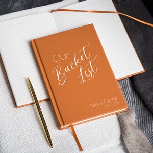 Puede incluir: Un diario de cuero marr&oacute;n con las palabras "Our Bucket List" en escritura blanca en la portada. Los nombres "Tara & Dennis" y la fecha "10-13-23" tambi&eacute;n est&aacute;n impresos en blanco en la portada.