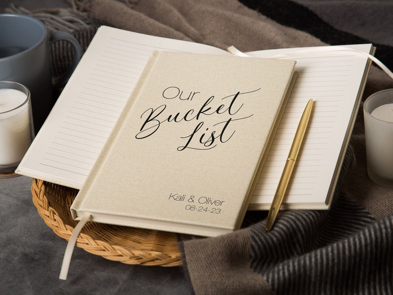 Puede incluir: Un diario beige cubierto de lino con el texto "Our Bucket List" en escritura cursiva negra. El diario est&aacute; abierto para revelar p&aacute;ginas rayadas y un bol&iacute;grafo dorado. El diario est&aacute; personalizado con los nombres "Kali & Oliver" y la fecha "08-24-23".