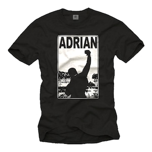Può includere: Maglietta nera con una grafica bianca di un uomo che alza il pugno in aria. La grafica include la parola "ADRIAN" in lettere bianche.