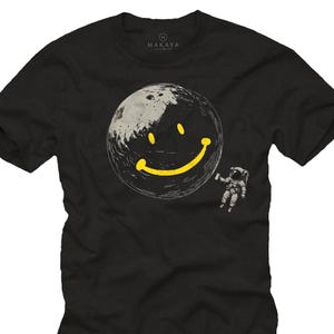 Puede incluir: Camiseta negra con una gran luna sonriente amarilla y gris. Un astronauta flota cerca de la luna. La palabra "MAKAYA" está impresa en la parte superior. Un diseño peculiar e imaginativo.