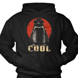 Könnte beinhalten: Schwarzer Kapuzenpullover mit einem Pinguin, der eine Sonnenbrille trägt, vor einem roten Kreis. Das Wort "COOL" ist darunter in einem Vintage-Stil gedruckt. Der Hoodie hat eine Vordertasche und eine Kapuze.