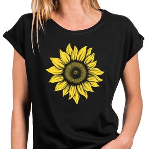 Può includere: T-shirt nero con un grande motivo a girasole giallo. Il girasole ha un centro dettagliato e petali giallo brillante. La maglietta ha maniche arrotolate e scollo tondo. La persona indossa jeans blu.