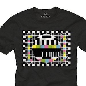 Puede incluir: Camiseta negra con un gráfico de patrón de prueba de televisión colorido. El gráfico presenta un fondo a cuadros blanco y negro con un círculo que contiene barras de colores y un rectángulo negro.