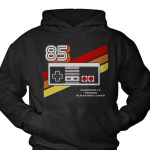 Può includere: Felpa con cappuccio nera con una grafica retrò di un controller per videogiochi e il numero 85. Il testo "Classically Trained Old School Gamer" è stampato sotto il controller.