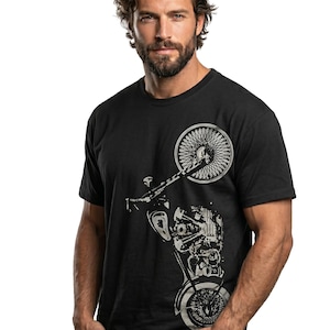 以下が含まれることがあります： 黒いTシャツに銀色のオートバイのグラフィック。デザインは、前輪、エンジン、その他の機械部品を示しています。Tシャツはクルーネックです。