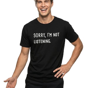 May include: Black crew neck t-shirt with the text "SORRY, I'M NOT LISTENING." printed in white. The person is wearing black pants and is smiling. The background is white.