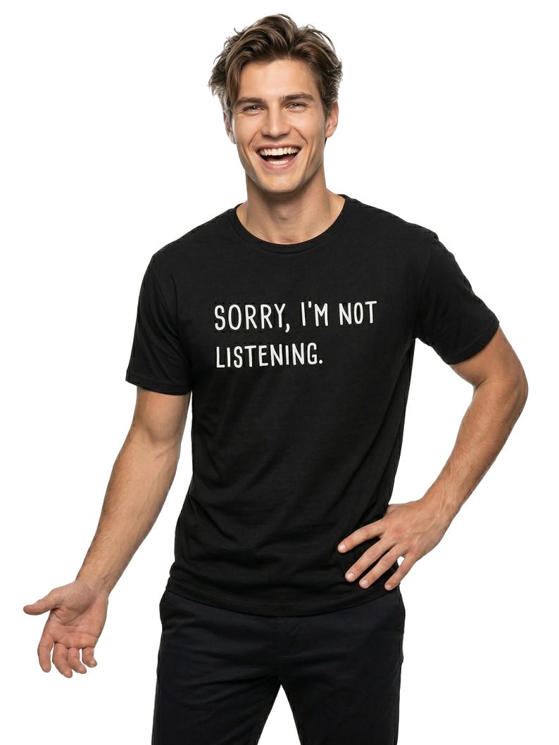 May include: Black crew neck t-shirt with the text "SORRY, I'M NOT LISTENING." printed in white. The person is wearing black pants and is smiling. The background is white.
