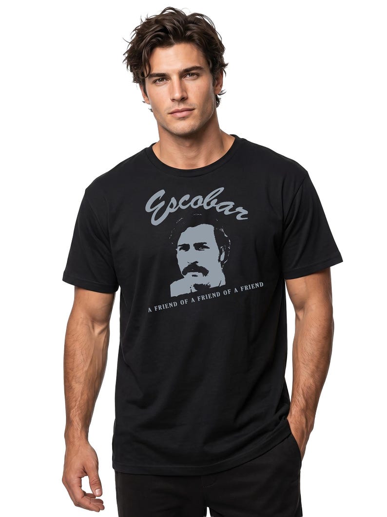 May include: Black t-shirt featuring a gray portrait and the name "Escobar". Below the portrait, the text reads "A FRIEND OF A FRIEND OF A FRIEND". The shirt has short sleeves and a crew neck.